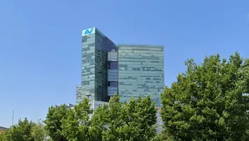 Modernes Bürogebäude mit Glasfassade und einem Logo auf dem Dach, umgeben von grünen Bäumen unter blauem Himmel.
