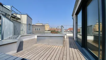 Geräumige Dachterrasse mit Holzboden und Blick über die Dächer der Stadt unter blauem Himmel.