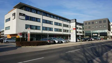 Modernes Geschäftsgebäude mit mehreren Etagen, großen Fenstern und Parkplätzen davor.