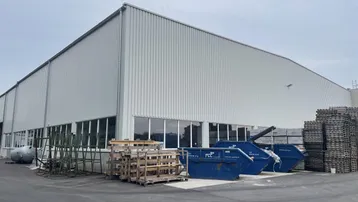 Großes Industriegebäude mit Wellblechfassade und breiten Fenstern, umgeben von einem gepflasterten Außenbereich mit Materiallagerung.