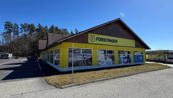 Gelbes Geschäftsgebäude mit großem Firmenlogo und Parkplätzen unter blauem Himmel an einem sonnigen Tag.