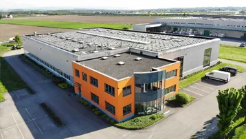 Luftaufnahme eines modernen Industriegebäudes mit orangefarbenem Bürotrakt und weitläufigen Parkplätzen.