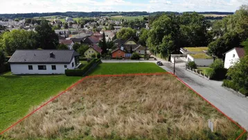 Luftaufnahme eines Baugrundstücks mit roter Umrandung, umgeben von grüner Landschaft und Wohnhäusern.