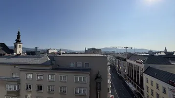 Panoramablick über die Stadt mit historischen Gebäuden, einer belebten Straße und fernen Bergen unter blauem Himmel.