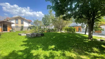 Sonniger Gartenbereich mit Rasenfläche, Bäumen und einem Hochbeet, umgeben von Wohnhäusern.