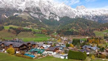 Panoramablick auf ein charmantes Dorf in den Alpen mit schneebedeckten Bergen im Hintergrund und grünen Hügeln.