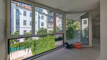 Unfertiger Innenraum mit großen Fenstern und Blick auf die umliegenden Gebäude und viel Grün.