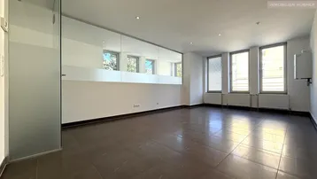 Heller, leerer Raum mit großen Fenstern, Fliesenboden und einer Glaswand.