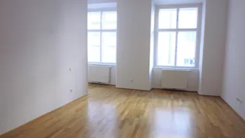 Heller, leerer Raum mit Holzboden und großen Fenstern, der viel Tageslicht hereinlässt.