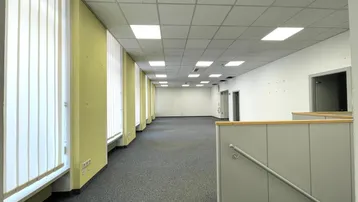Geräumiges, leeres Büro mit Teppichboden, Rasterdecke und vertikalen Jalousien an den Fenstern.
