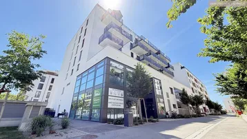 Modernes Gebäude mit Glasfassade und Balkonen unter blauem Himmel, umgeben von Bäumen.