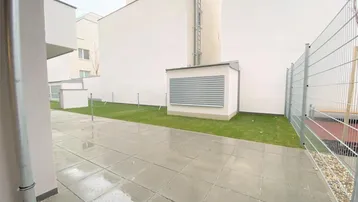 Gepflasterte Terrasse mit angrenzender Rasenfläche und modernen Gebäuden im Hintergrund.