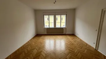 Helles, leeres Zimmer mit Fischgrätparkettboden und Blick auf die gegenüberliegenden Gebäude.
