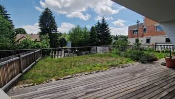 Geräumige Holzterrasse mit Blick ins Grüne und einem kleinen Rasenbereich unter blauem Himmel.