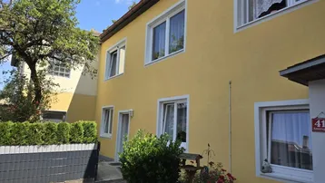 Gelbe Hausfassade mit weißen Fenstern und einem kleinen Vorgartenbereich unter blauem Himmel.