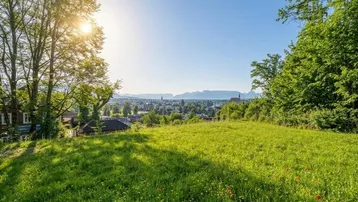 Weitläufige Grünfläche mit Blick auf die Stadt und die umliegende Berglandschaft unter strahlend blauem Himmel.