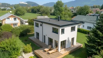 Modernes weißes Haus mit Flachdach und großer Holzterrasse, umgeben von Grünflächen und Bergblick.