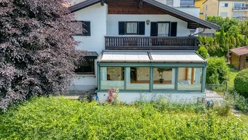 Hausfassade mit Wintergarten, Balkon und üppiger grüner Umgebung unter bewölktem Himmel.
