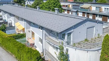 Mehrfamilienhaus mit grauen Dächern, weißen Fassaden und grünen Hecken in bergiger Umgebung.