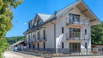 Modernes Mehrfamilienhaus mit mehreren Balkonen und Satteldach in grüner Umgebung.