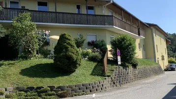 Gelbes Mehrfamilienhaus mit Satteldach, Balkonen und gepflegtem Vorgarten an einer Straße.