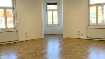 Geräumiges, leeres Zimmer mit Fischgrätparkett, hohen Fenstern und viel Tageslicht.