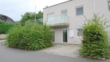 Modernes Gebäude mit Balkon und gepflastertem Vorplatz, umgeben von grünen Hecken.
