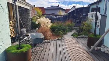 Gestalteter Gartenbereich mit Holzterrasse, Pflanzen und Brennholzstapel unter einem Unterstand.