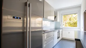Funktionale Küche mit weißen Hochglanzschränken, großem Siemens Kühlschrank und Fenster mit Tageslicht.