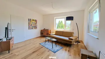 Helles Wohnzimmer mit Holzboden, beigem Sofa und Blick ins Grüne durch große Fenster.