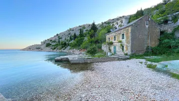 Altes Steinhaus direkt am Kiesstrand einer ruhigen Bucht mit klarem Wasser und grünen Hügeln.
