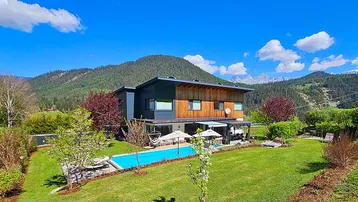 Modernes Haus mit großem Garten, Pool und Bergblick unter blauem Himmel.