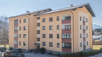 Mehrfamilienhaus mit mehreren Etagen, Balkonen und Parkplätzen im Freien unter blauem Himmel.