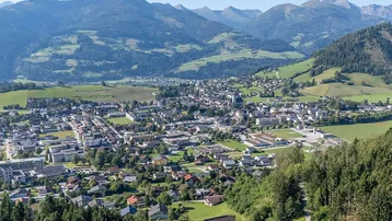 Panoramablick auf eine malerische Stadt in einem Tal, umgeben von grünen Bergen und Wäldern.