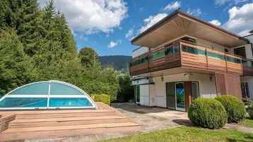 Außenansicht des Hauses mit überdachtem Poolbereich und Holzterrasse, umgeben von Bäumen.