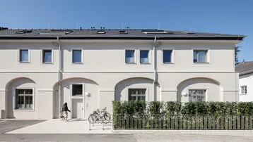 Modernes Mehrfamilienhaus mit heller Fassade, großen Fenstern und gepflegter Hecke.