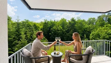 Ein Paar genießt den Ausblick von einem möblierten Balkon mit Blick auf grüne Bäume.
