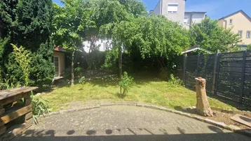 Grüner Gartenbereich mit Rasenfläche, Bäumen und einer kleinen gepflasterten Terrasse im Sonnenschein.