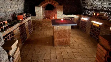 Historischer Weinkeller mit Ziegelgewölbe, Flaschenregalen und zentralem Steintisch, stimmungsvoll beleuchtet.
