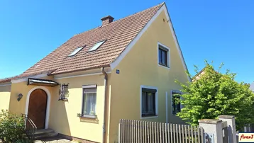 Gelbes Einfamilienhaus mit Satteldach, Dachfenstern und Holzzaun im Vorgarten unter blauem Himmel.
