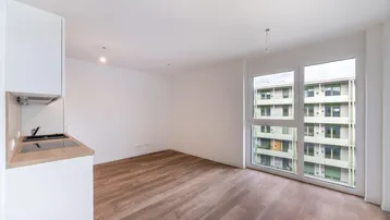 Heller Wohnbereich mit integrierter Küchenzeile, Holzboden und großen Fenstern mit Blick auf ein Gebäude.