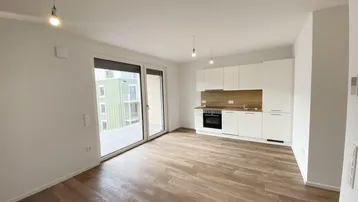 Heller offener Wohnbereich mit integrierter Küche, modernem Laminatboden und Zugang zum Balkon.