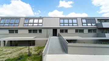 Moderne Gebäudefassade mit Rampe zum Eingang und Dachfenstern unter blauem Himmel.