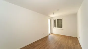 Helles, leeres Zimmer mit Holzfußboden und Zugang zu einem Balkon oder einer Terrasse.