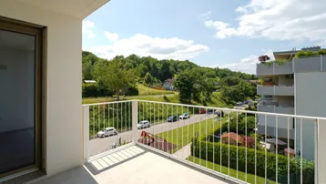 Geräumiger Balkon mit weißem Geländer und weitem Blick über eine Straße, Spielplatz und grüne Hügel.