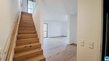 Helles Treppenhaus mit Holzstufen und angrenzendem, leeren Wohnbereich mit Parkettboden.