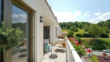 Sonniger Balkon mit modernen Sitzgelegenheiten, Blumenkästen und weitem Blick ins Grüne sowie auf einen Spielplatz.