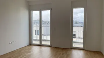 Heller, unmöblierter Raum mit Holzboden und großen Fenstern, die auf einen französischen Balkon führen.
