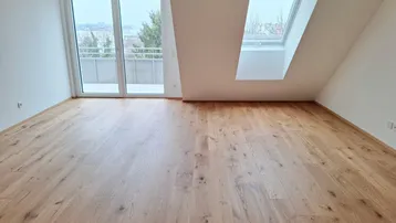 Heller Wohnraum im Dachgeschoss mit Holzboden, großem Fenster und Zugang zum Balkon.