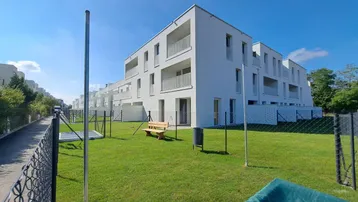 Moderne Wohngebäude mit weißer Fassade und Balkonen, umgeben von einer gepflegten Grünfläche unter blauem Himmel.